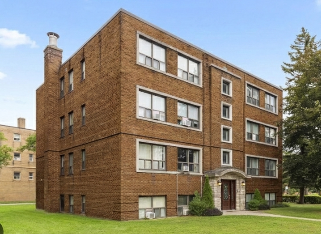 A four-story apartment building with a dark brown brick façade. The building features a central entrance with a stone portico and arched doorway, large rectangular windows with white frames, and a chimney on the left. A green lawn spans the foreground, with a large tree visible on the right side.